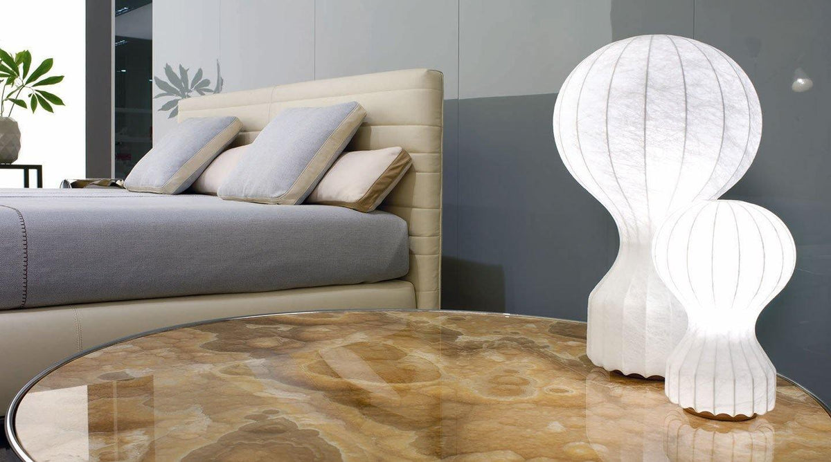 Two Clérachic lamps lit on a glass-topped round table in front of a bed with grey cushions.
