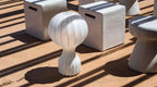 Clérachic lamp outdoors on a sunlit terracotta-coloured terrace, with striped shadows and pale concrete stools around it.