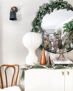 Tall Clérachic lamp on a white cabinet at Christmas, beside a eucalyptus wreath, a round mirror and a copper cone.