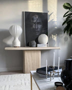 A Clérachic table lamp on a travertine console with a black-and-white portrait print, a stone sphere and tall candles.