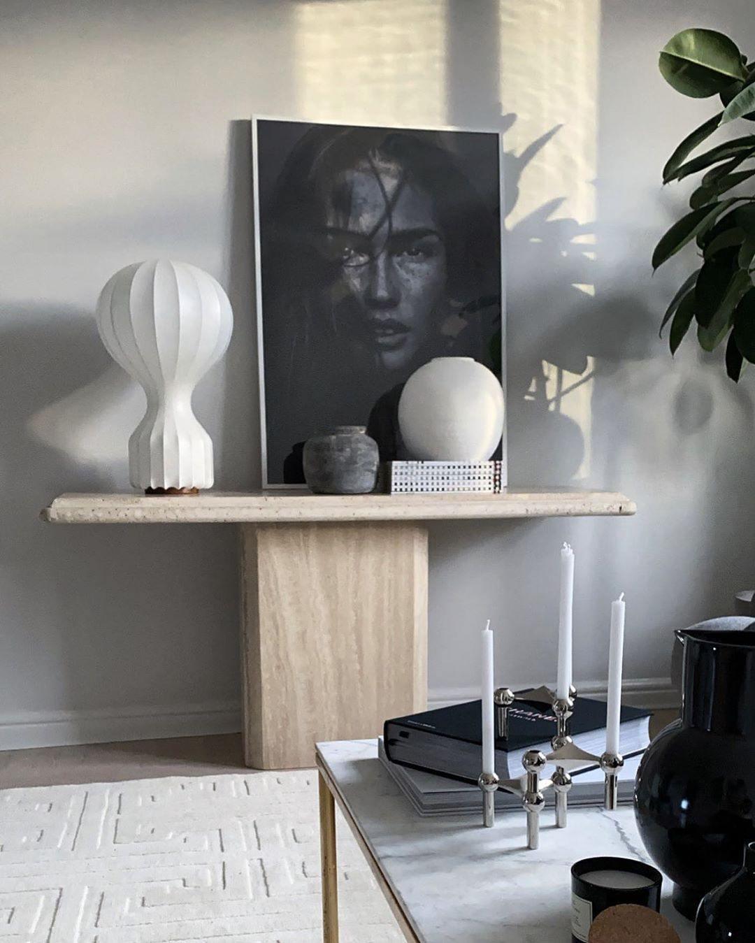A Clérachic table lamp on a travertine console with a black-and-white portrait print, a stone sphere and tall candles.