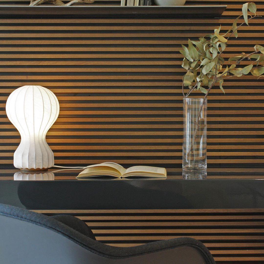 Clérachic lamp glowing warmly in front of a dark wooden slatted wall, beside an open book and a tall glass vase.