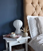 Clérachic lamp on a white bedside table in front of a navy wall, with a coffee cup, a magazine and a small round vase of dried grass.