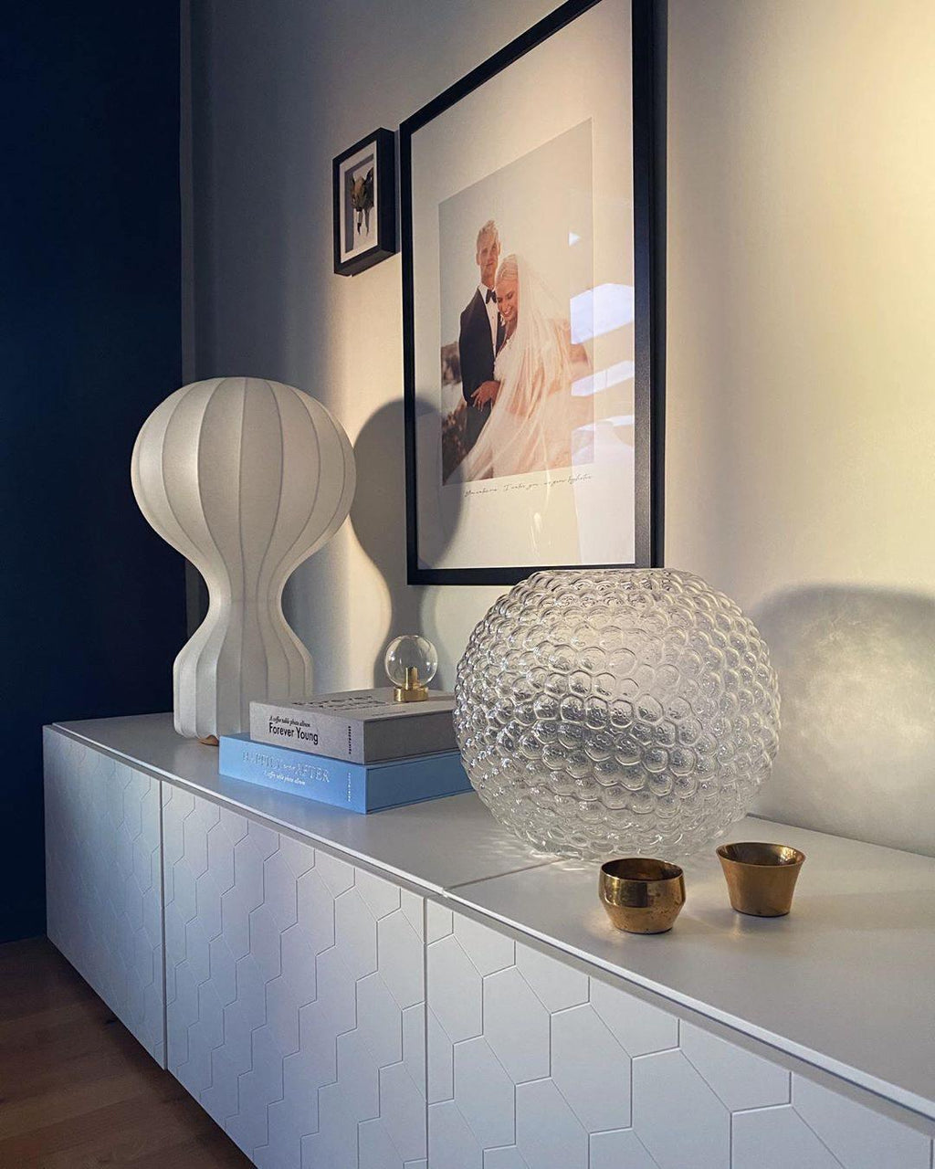 A Clérachic lamp on a white sideboard under a framed portrait, with a faceted glass globe and two small brass cups.