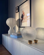 A Clérachic lamp on a white sideboard under a framed portrait, with a faceted glass globe and two small brass cups.