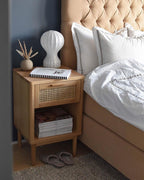 Clérachic table lamp on a rattan-fronted bedside table, beside a pillowed bed and a dark blue wall.