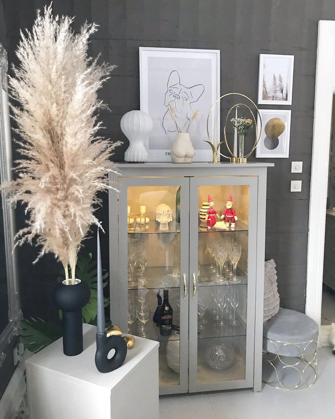 Small Clérachic lamp on top of a glazed grey display cabinet, with framed prints, a tall black vase and pampas grass beside it.