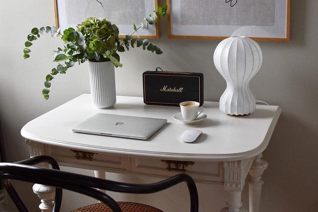 A Clérachic lamp on a white writing desk next to a laptop, a coffee cup and a retro speaker, below two framed prints.