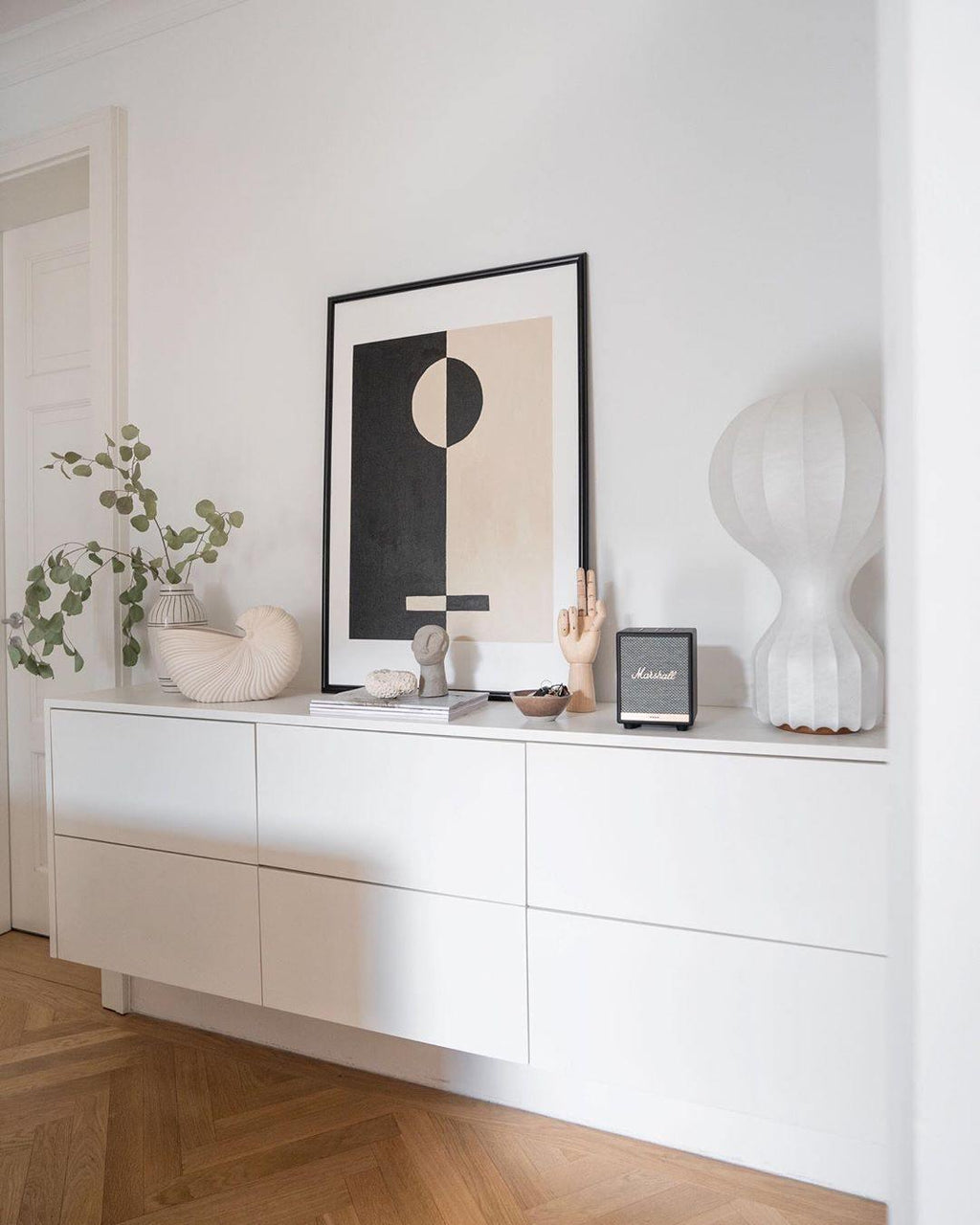 Clérachic table lamp at the end of a white wall-hung sideboard, with a framed graphic print, a shell and a small speaker.