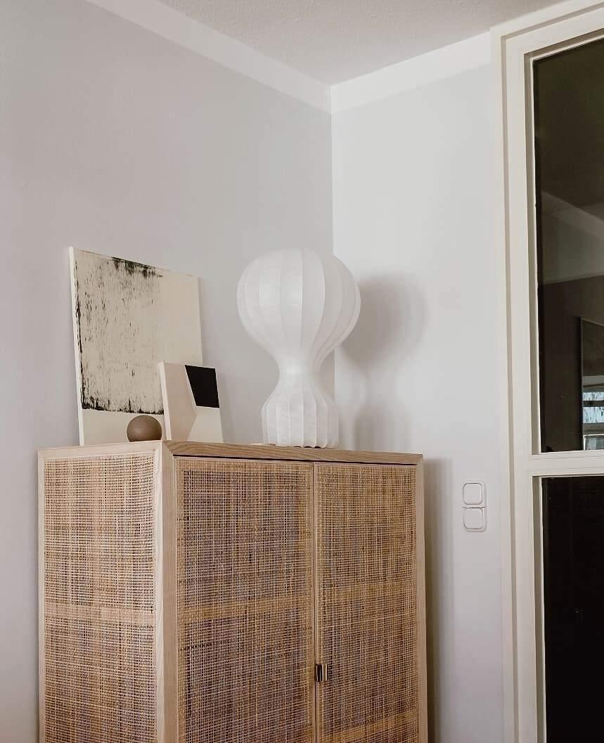 A Clérachic lamp standing on a rattan cabinet in a corner, in front of an abstract canvas.