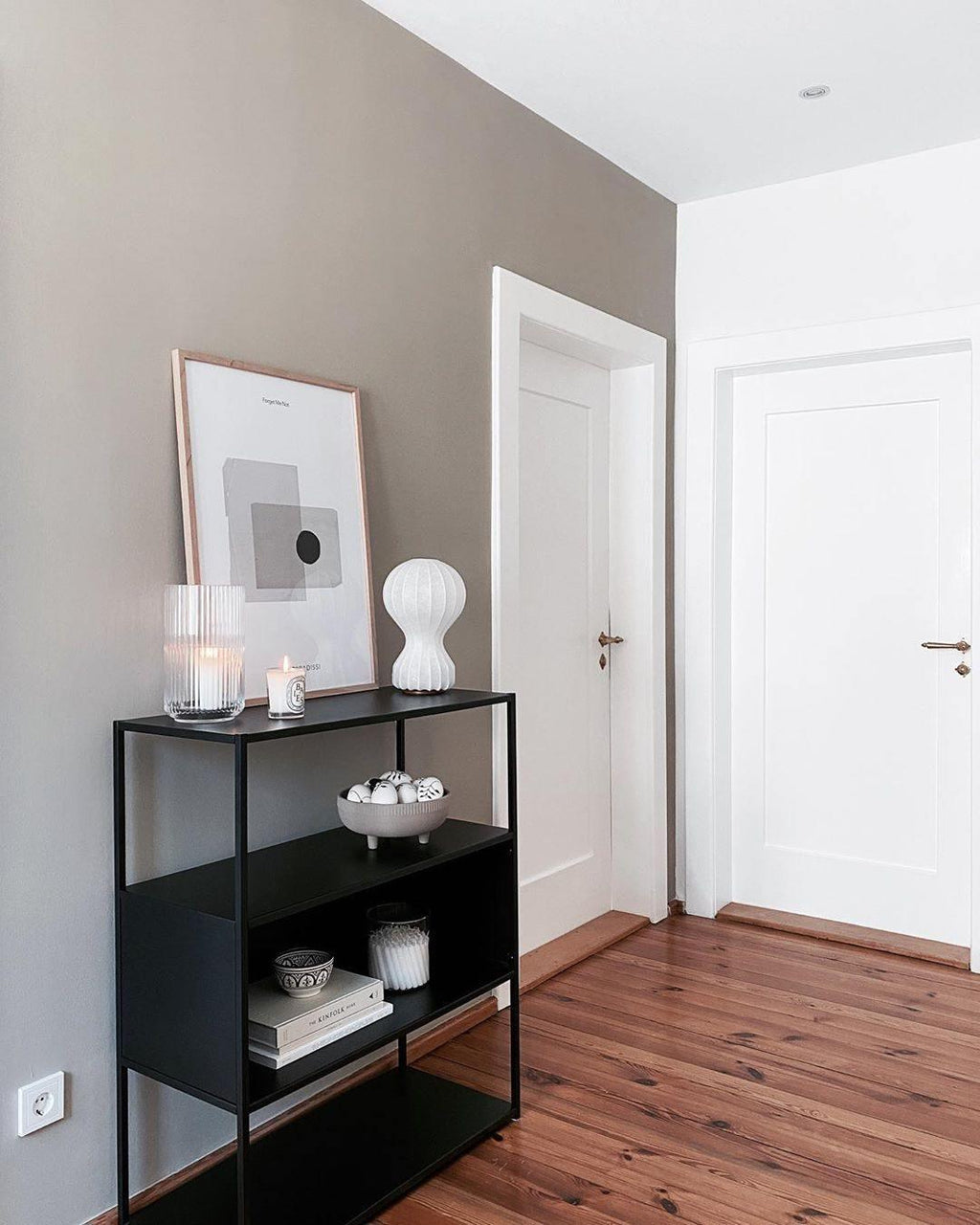 Small Clérachic lamp on a black metal shelving unit in a hallway, beside a framed print and a bowl of decor.