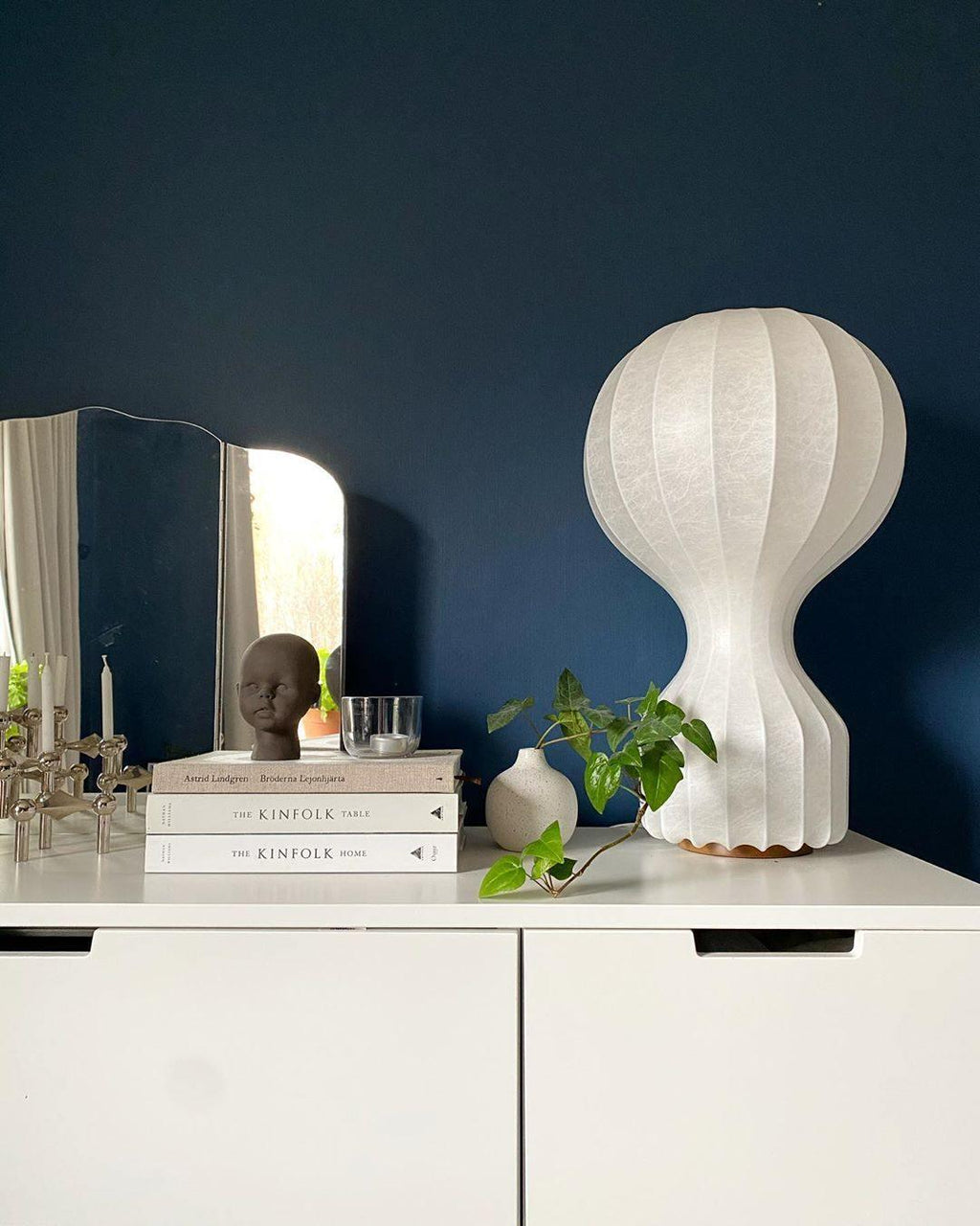 White Clérachic lamp on a white chest of drawers in front of a deep navy wall, with books, a vase and a small mirror.