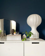 White Clérachic lamp on a white chest of drawers in front of a deep navy wall, with books, a vase and a small mirror.