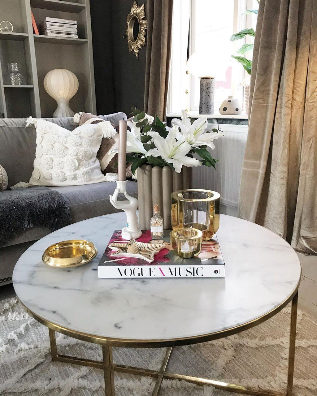 A Clérachic lamp on a shelf in the background of a living room, behind a round marble coffee table with brass edges.