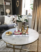A Clérachic lamp on a shelf in the background of a living room, behind a round marble coffee table with brass edges.