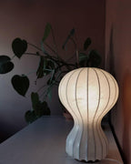 Close view of a lit Clérachic lamp glowing amber on a dark surface, with a leafy houseplant against a dusky rose wall.