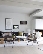 A small Clérachic lamp across a white living room, by the fireplace, with cane chairs and a pale round coffee table.