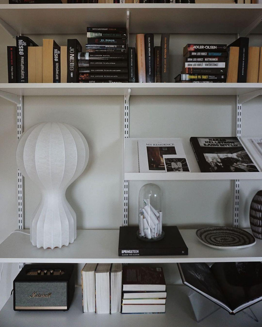 Clérachic table lamp on an open white shelf among rows of books, a glass cloche and an old speaker.