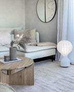 Lit Clérachic lamp on the floor beside a pale sofa and a round oak coffee table, against a plaster-grey wall.
