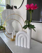 Partial view of a Clérachic lamp behind two brass hoops, a white arched vase and a pink peony on a white console.