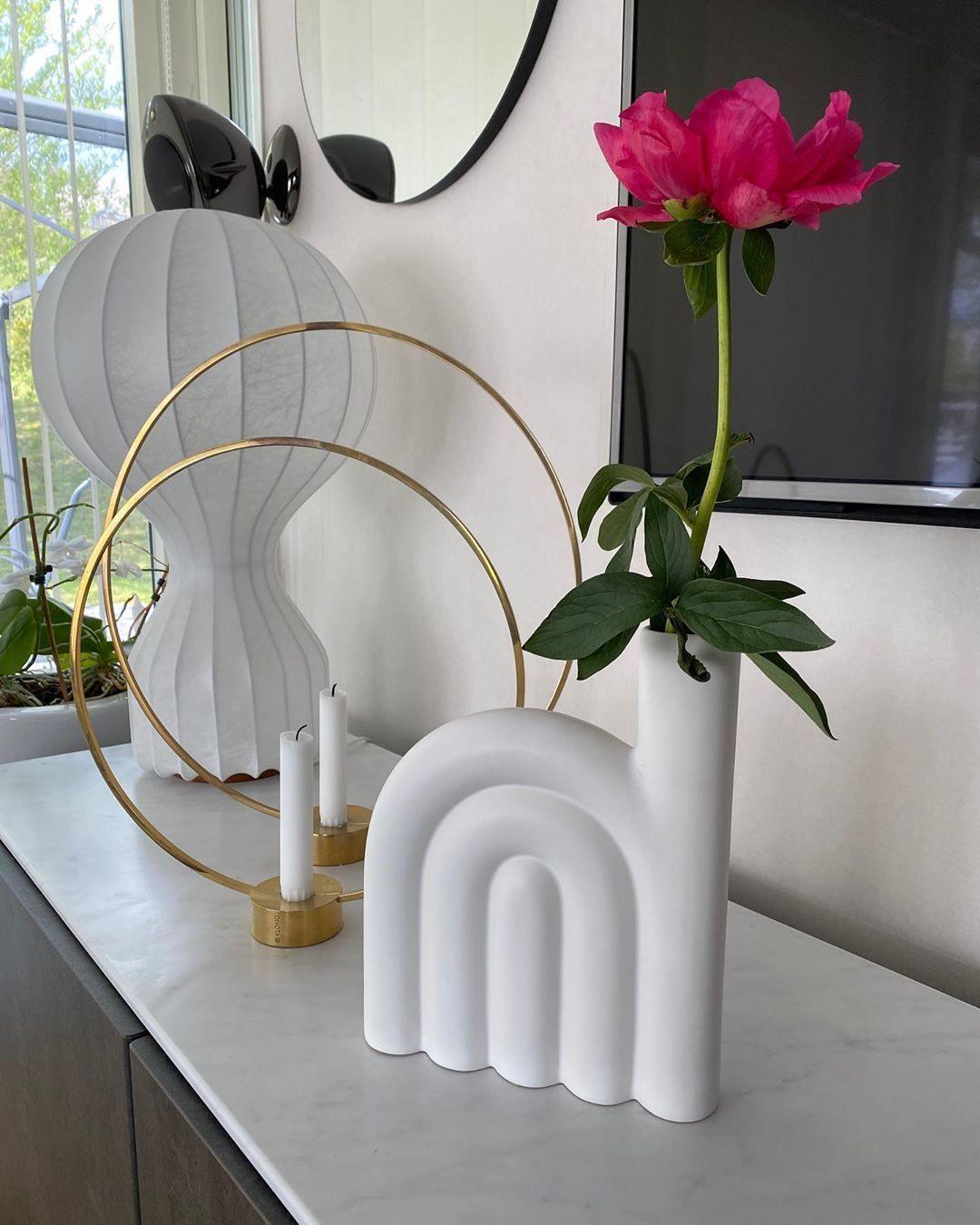 Partial view of a Clérachic lamp behind two brass hoops, a white arched vase and a pink peony on a white console.