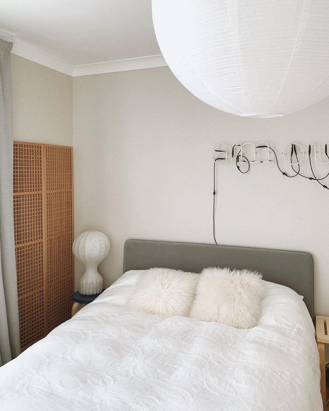Small Clérachic lamp beside a single bed with white fluffy cushions, below a large white paper pendant.