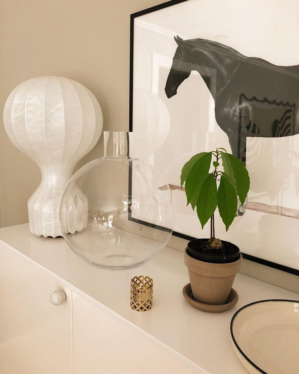 Clérachic table lamp on a cream cabinet in front of a large print of a dark horse, with a glass vase and a potted plant.