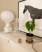 Clérachic table lamp on a cream cabinet in front of a large print of a dark horse, with a glass vase and a potted plant.