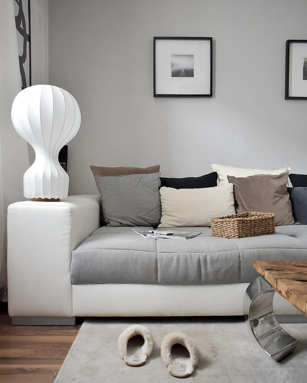 A Clérachic table lamp on the arm of a white sofa with grey cushions, above a pair of sheepskin slippers on the rug.