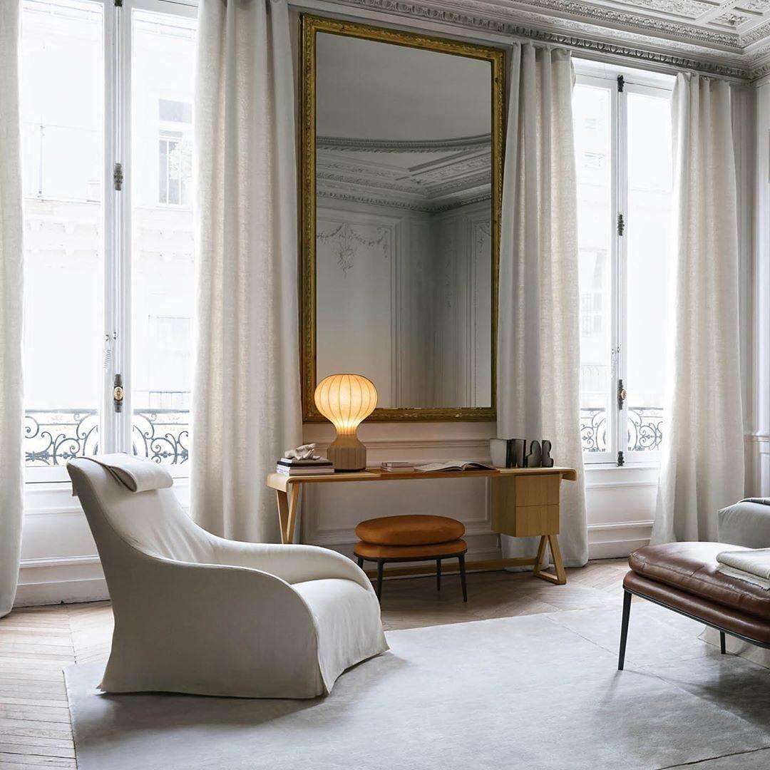 A Clérachic lamp glowing on a desk in a tall Parisian-style room, with a pale armchair and a gilt-framed mirror.