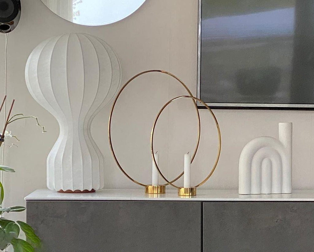 Close view of a larger Clérachic lamp on a grey cabinet, beside two brass rings with candles and a white arched ornament.