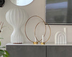 Close view of a larger Clérachic lamp on a grey cabinet, beside two brass rings with candles and a white arched ornament.