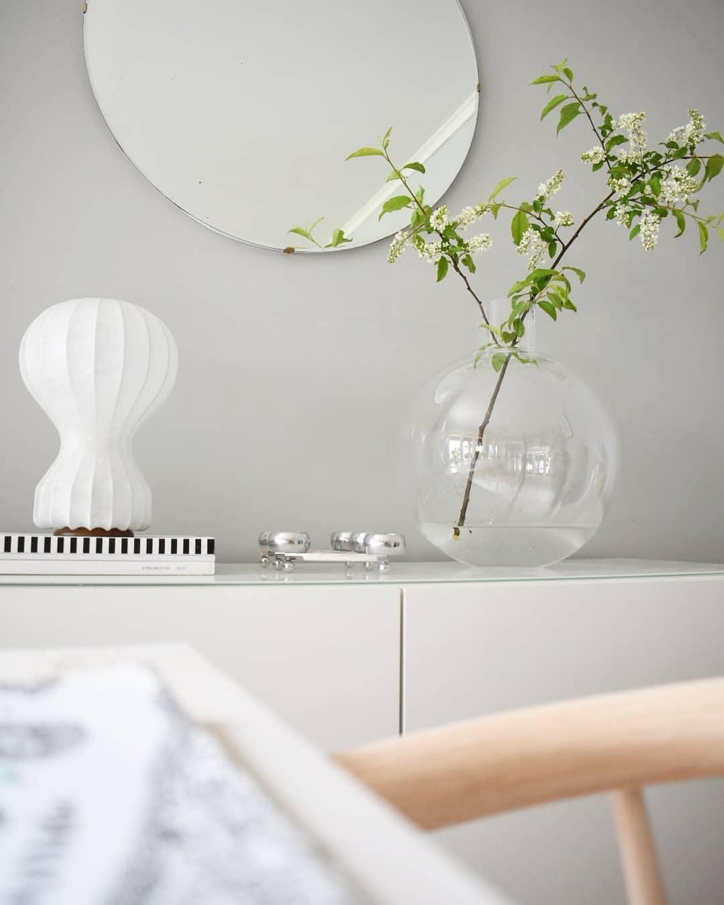Clérachic table lamp on a white sideboard under a round mirror, beside a glass globe vase with a flowering branch.