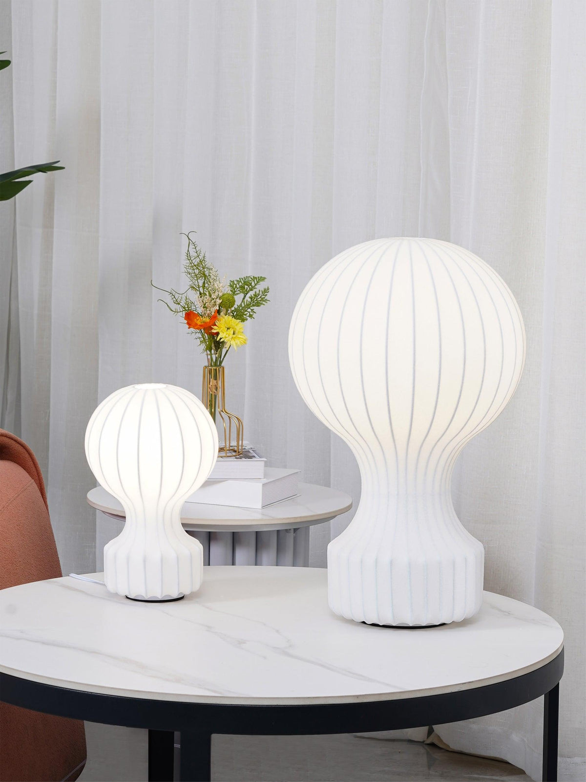 Both sizes of the Clérachic lamp lit on a round marble-topped table, with sheer curtains and a small vase of flowers behind.