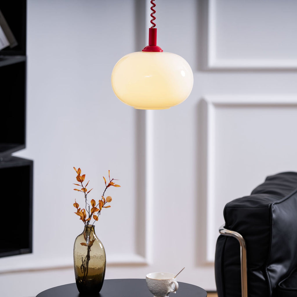 A Clérachic pendant with a red cap and coil cable glowing warm above a side table, a smoky glass vase and a black leather chair.