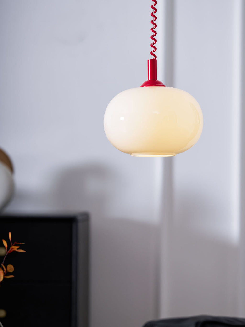 Red-capped Clérachic pendant hanging in front of a pale wall, its milky shade lit softly, with a black cabinet at the edge of the frame.