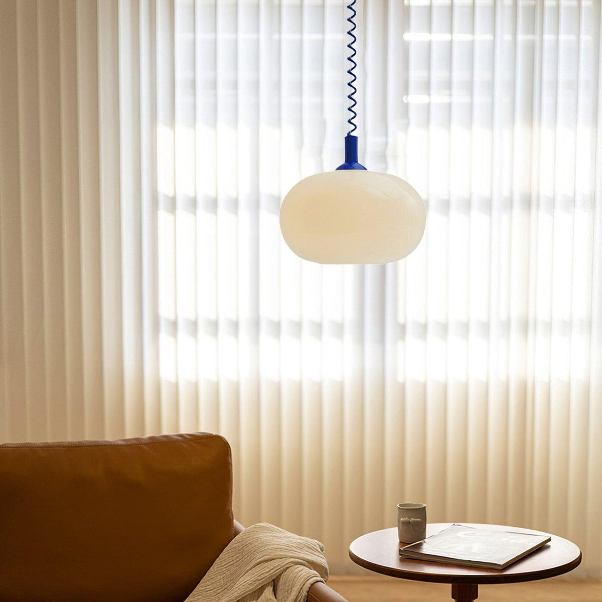 A blue-capped Clérachic pendant hung low in front of sheer vertical blinds, with a tan sofa and a small round table below.