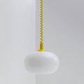 This Clérachic pendant with a yellow cap and yellow coil cable, its white glass shade unlit, against a pale grey wall corner.