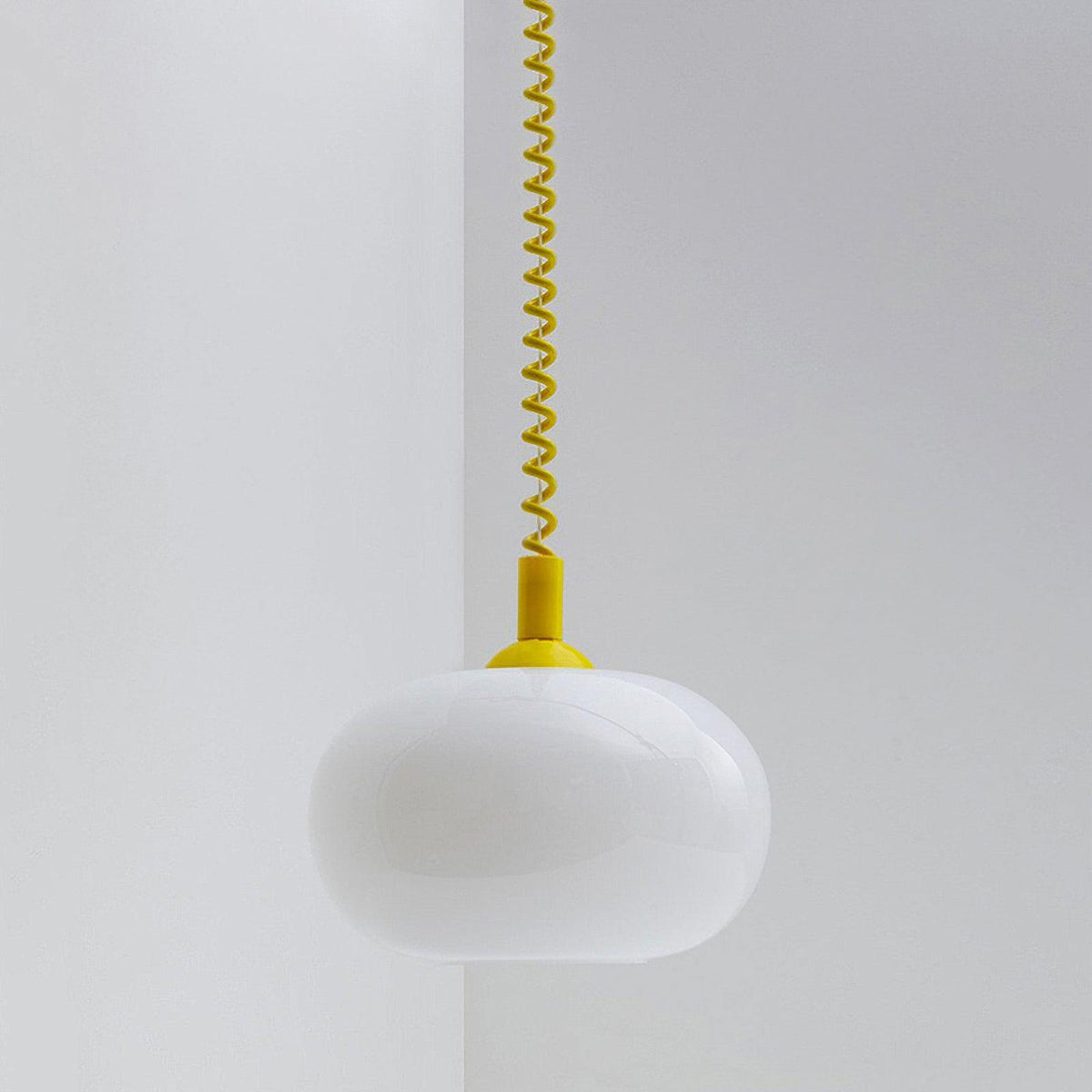 This Clérachic pendant with a yellow cap and yellow coil cable, its white glass shade unlit, against a pale grey wall corner.