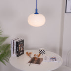 Blue-capped Clérachic pendant above a round white table with books, a checked mug and a fern, against a light wall.
