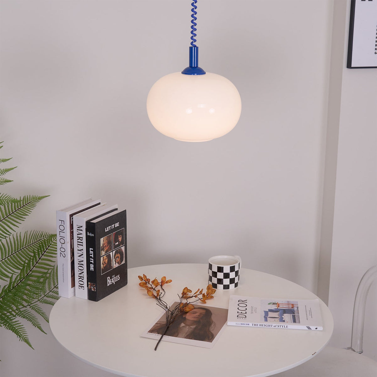 Blue-capped Clérachic pendant above a round white table with books, a checked mug and a fern, against a light wall.