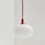 A Clérachic pendant with a red cap and red coil cable, its white glass shade unlit, hanging in a pale room corner.