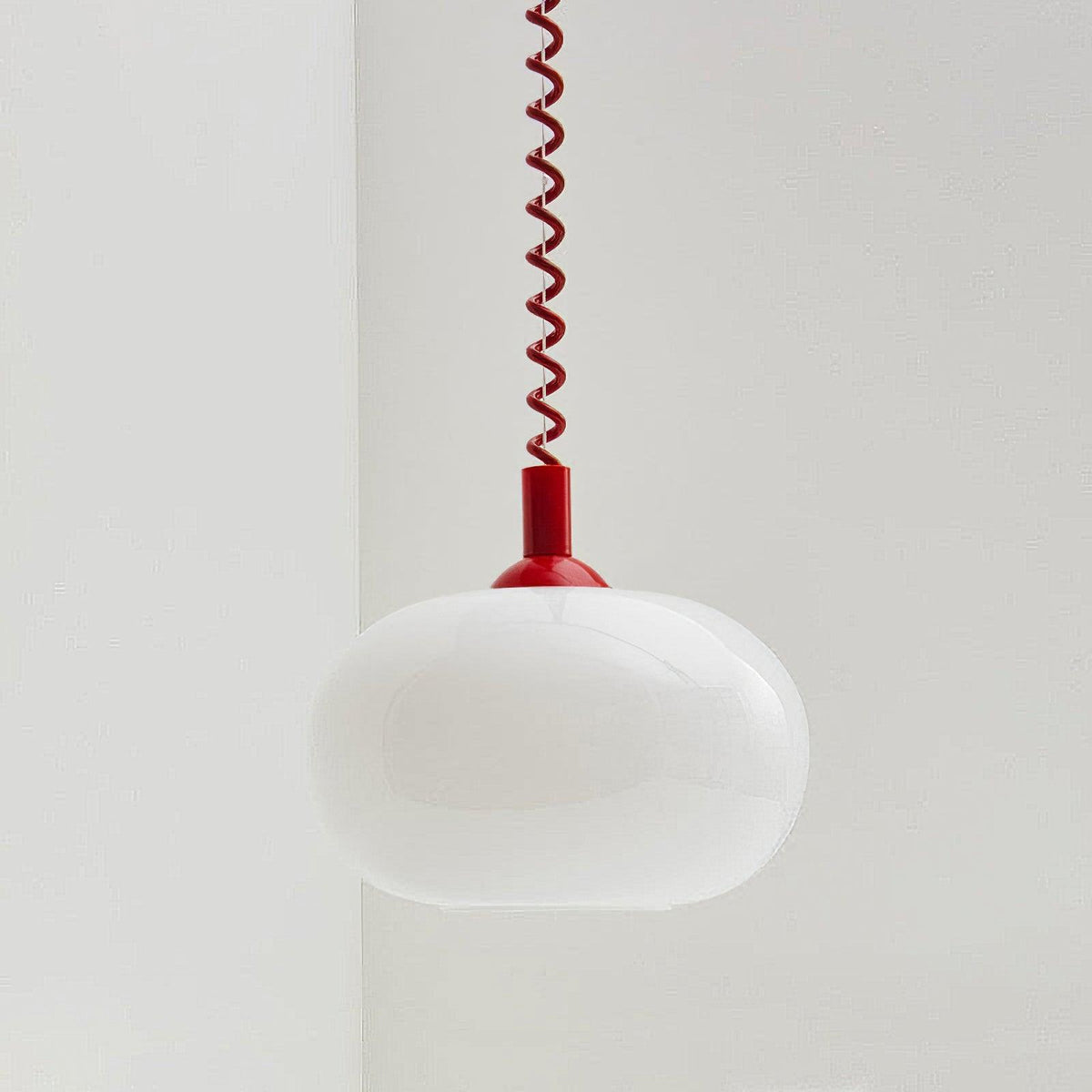 A Clérachic pendant with a red cap and red coil cable, its white glass shade unlit, hanging in a pale room corner.