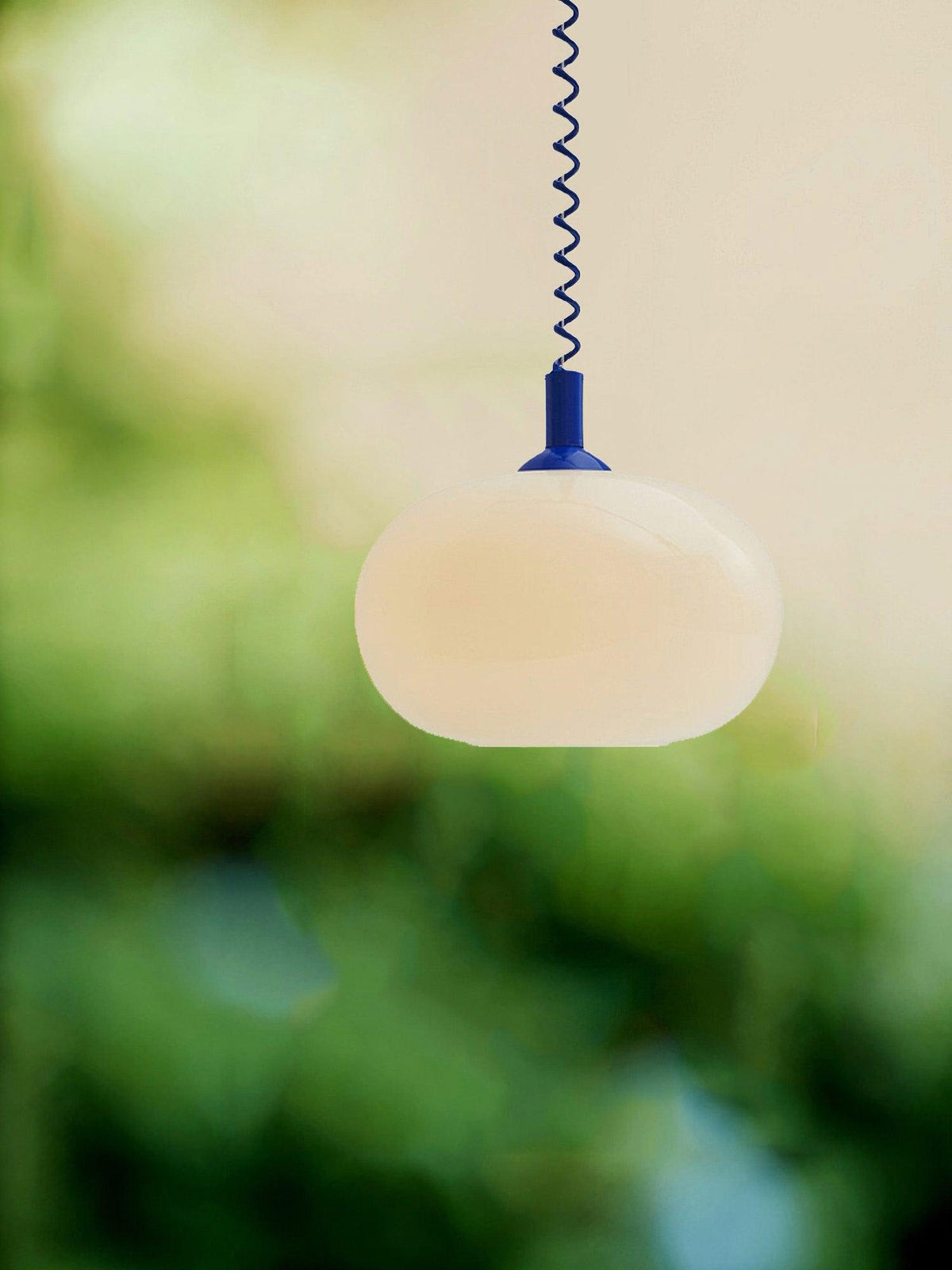 A lit Clérachic pendant with a blue cap and cable, its cream shade set against a soft green blurred garden.