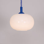 Close view of the lit blue-capped Clérachic pendant, its rounded shade glowing warm against a grey-mauve wall.