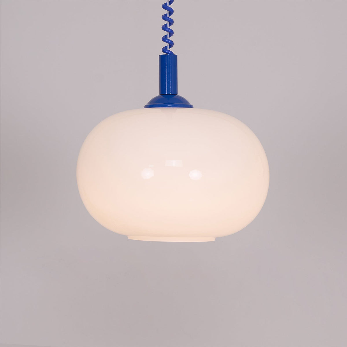 Close view of the lit blue-capped Clérachic pendant, its rounded shade glowing warm against a grey-mauve wall.