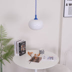 A small Clérachic pendant with a blue cap hung above a round white table with books, a checked mug and a fern.