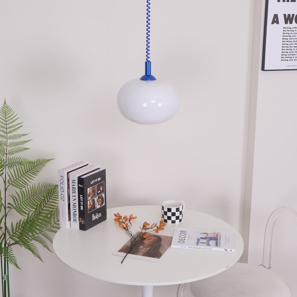 A small Clérachic pendant with a blue cap hung above a round white table with books, a checked mug and a fern.