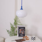 Blue-capped Clérachic pendant unlit above a white table with a fern, stacked books and a checked mug.