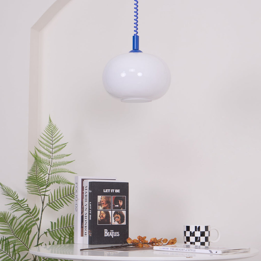 Unlit Clérachic pendant with a blue cap hanging beside a fern and a round white table with books and a mug.